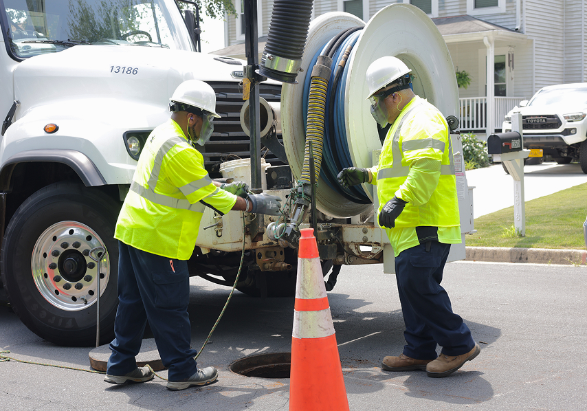 Two workers in safety gear operating equipment near an open manhole on a residential street